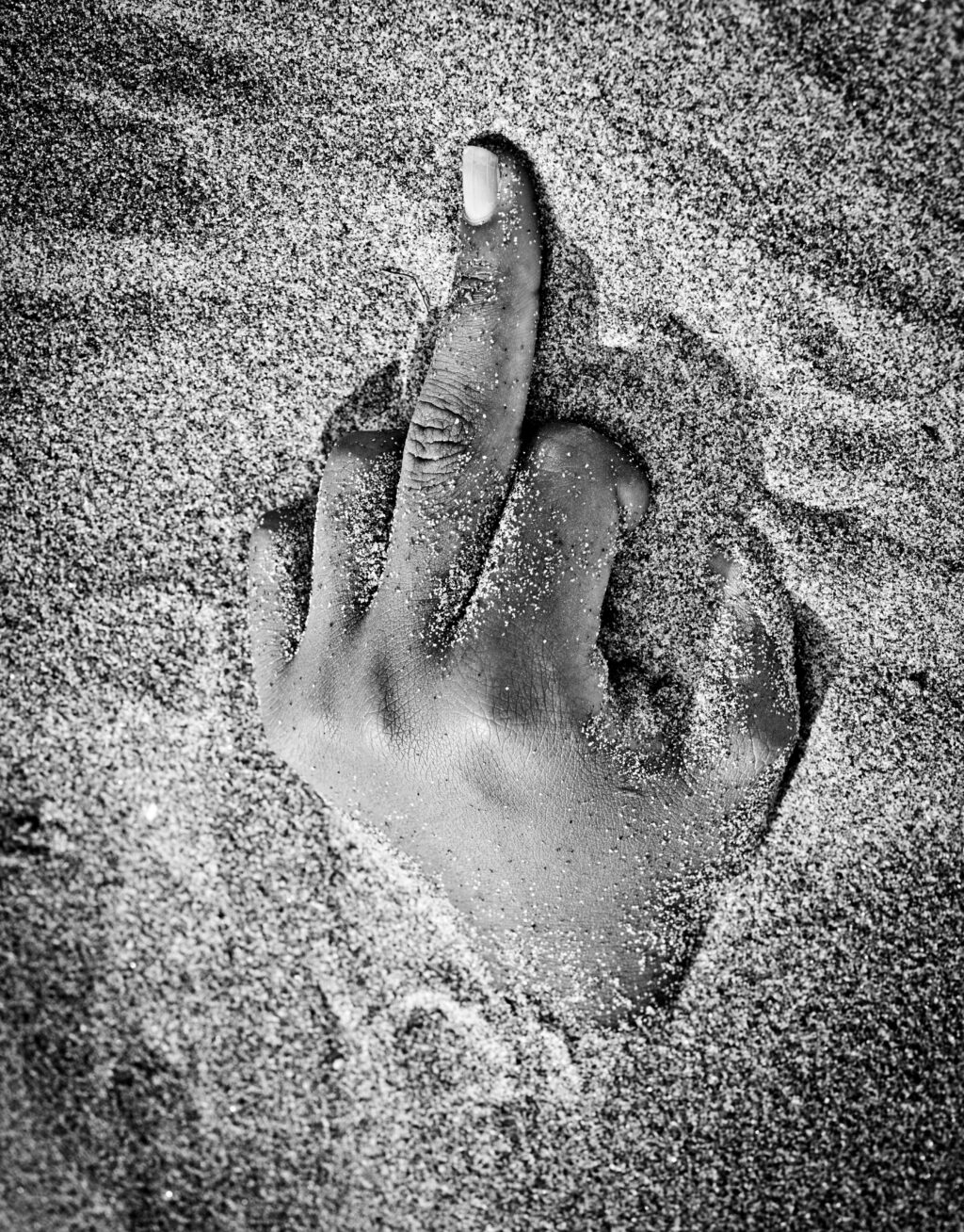 Mano che emerge dalla sabbia con il dito medio alzato, fotografia in bianco e nero